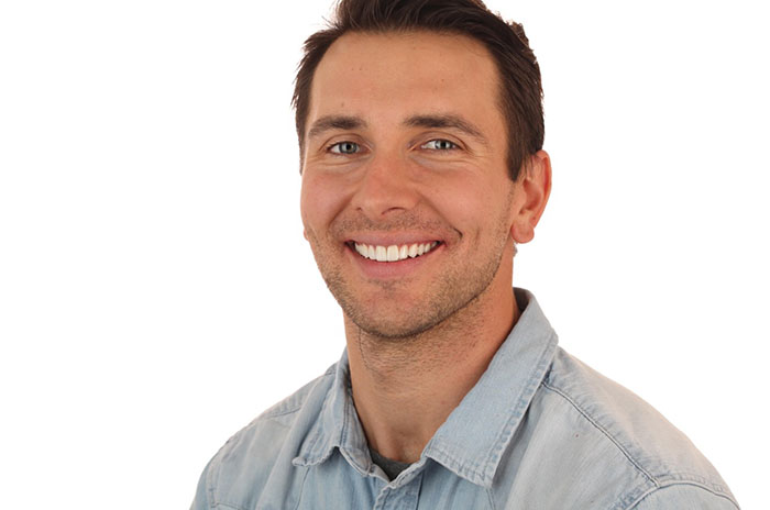 A smiling man with short hair wearing a blue denim shirt against a white background.