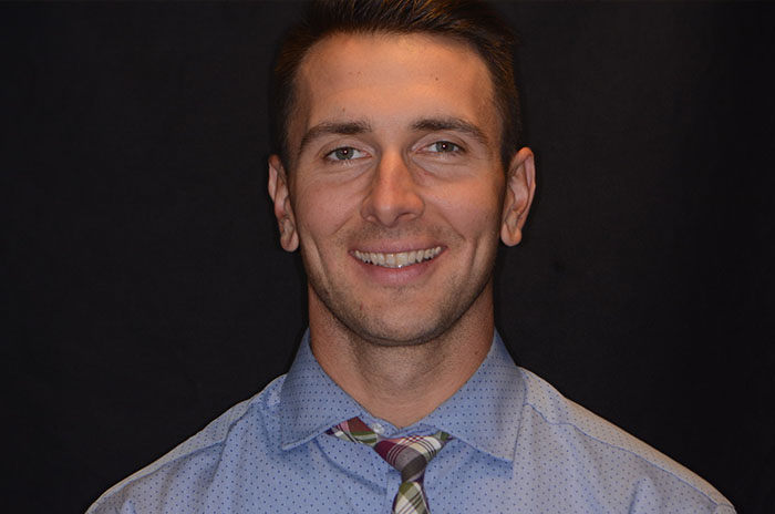 The image features a man smiling at the camera, wearing a patterned shirt, with short hair and a beard. He has a tattoo on his right arm.