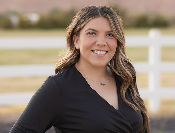 The image shows a woman smiling at the camera, standing outdoors in front of a fence with a white picket design, wearing a black top and posing for a portrait.