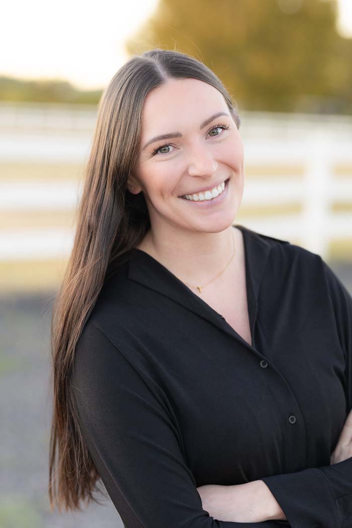A woman with long hair, smiling at the camera, wearing a dark top and standing outdoors against a blue sky background.
