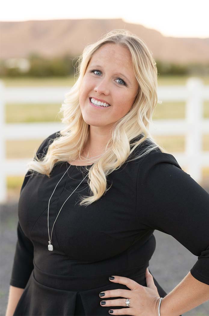 A woman wearing a black dress stands outdoors with a smile, posing for a portrait.