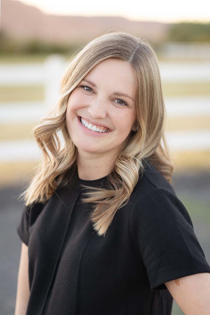 The image shows a smiling woman with blonde hair wearing a black top, posing for a portrait outdoors during daylight hours.