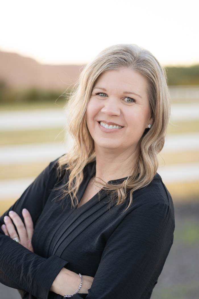 A woman standing outdoors with her arms crossed, wearing a black top, against a sunset backdrop.