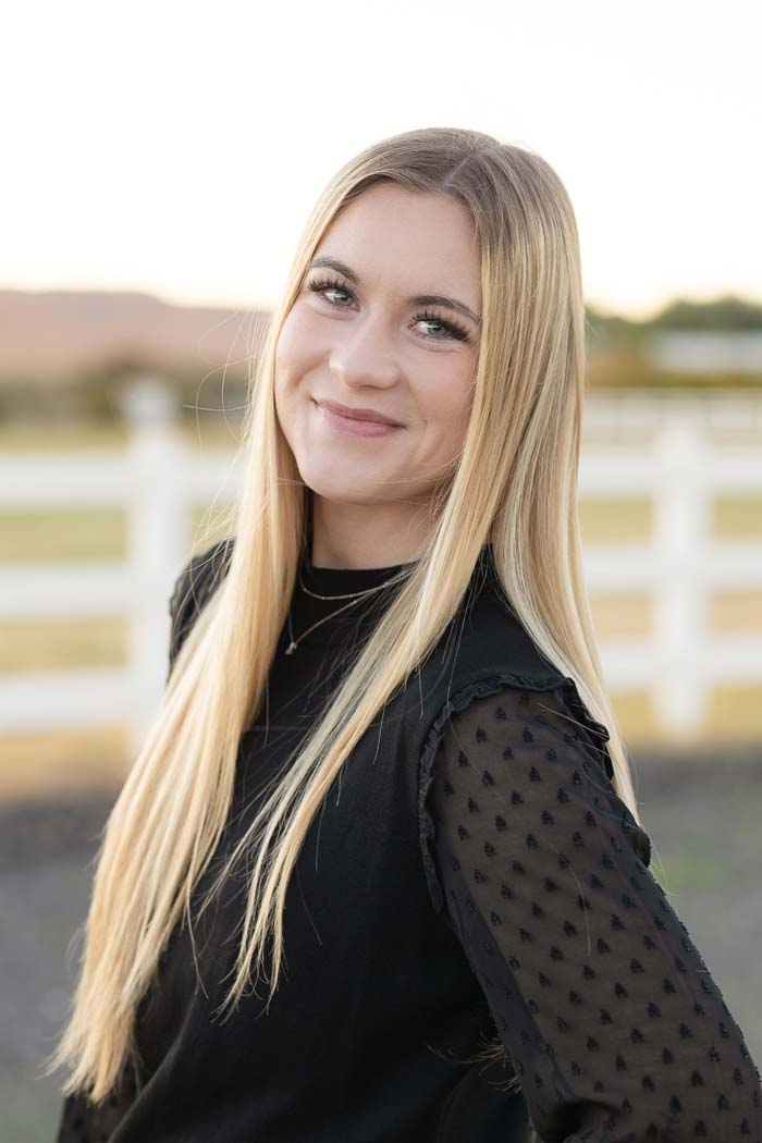 A young woman with long blonde hair, wearing makeup, smiling at the camera, standing outdoors in front of a fence during sunset.