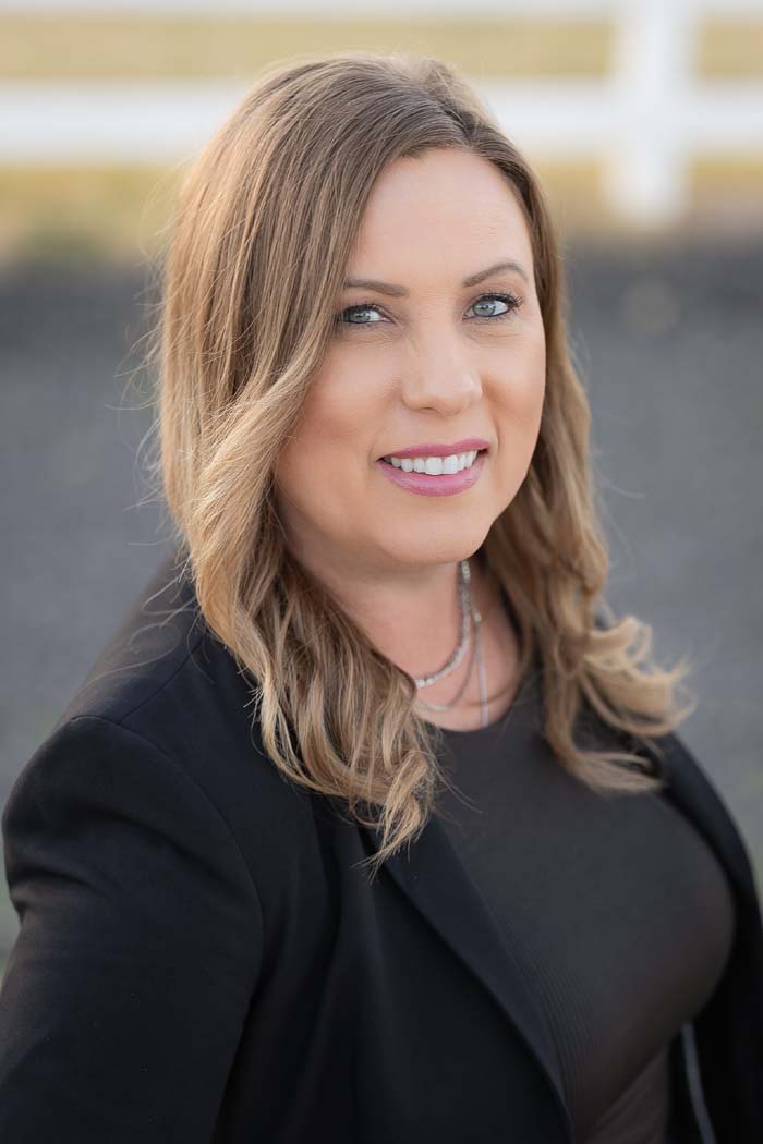 The image shows a woman wearing a black blazer and smiling at the camera. She has short hair and is standing outdoors with a clear sky in the background.