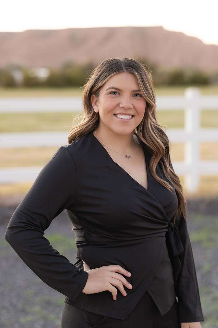 A woman in a black dress stands confidently outdoors with a white fence behind her.